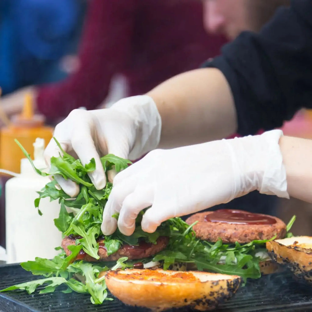 Ein chef trägt hygienische weiße nitrilhandschuhe und legt frischen rucola auf einen gegrillten burgerpatty