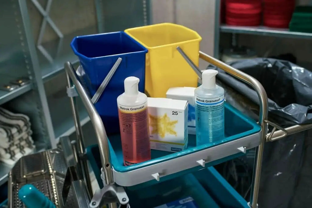 Blaue und gelbe kunststoffeimer mit metallgriffen auf einem wagen neben flaschen wetrok granusan sanitärreiniger sticks